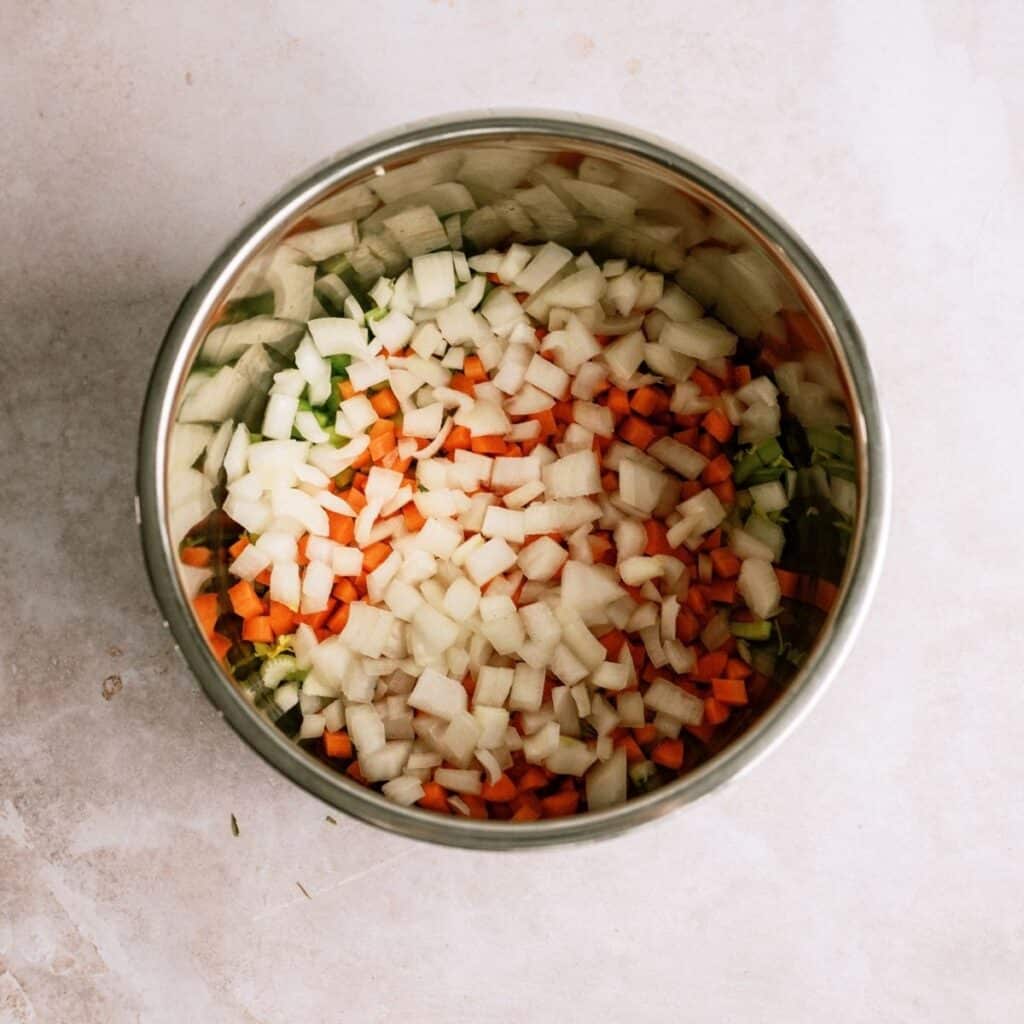 A metal bowl filled with chopped onions, carrots, and celery on a light-colored surface.
