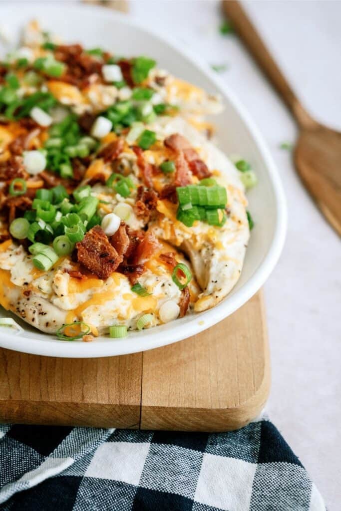 A white bowl filled with a baked potato dish topped with chopped green onions, bacon, and melted cheese sits on a wooden cutting board next to a blue and white checkered cloth.