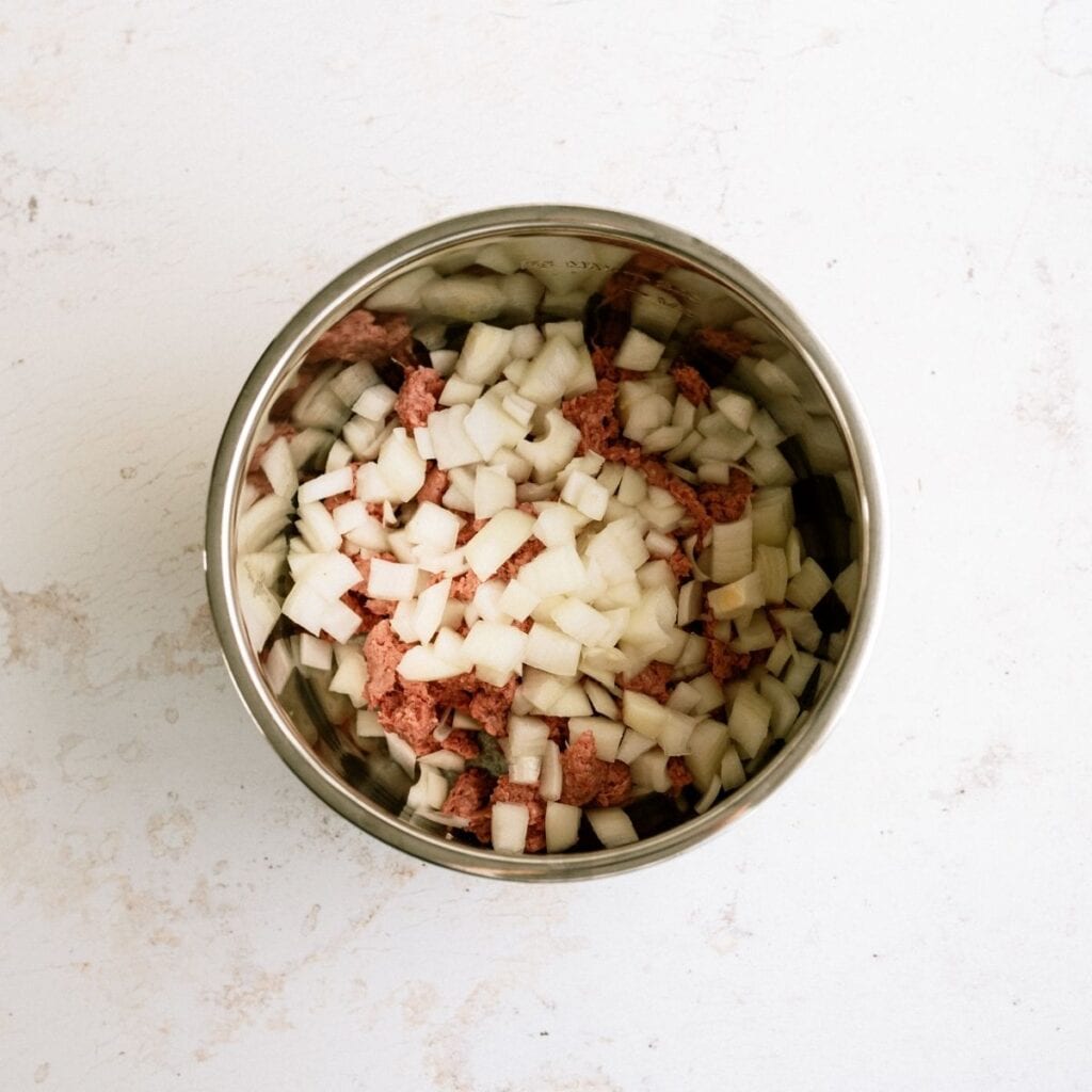 A metal bowl containing diced onions and raw ground meat on a light-colored surface.