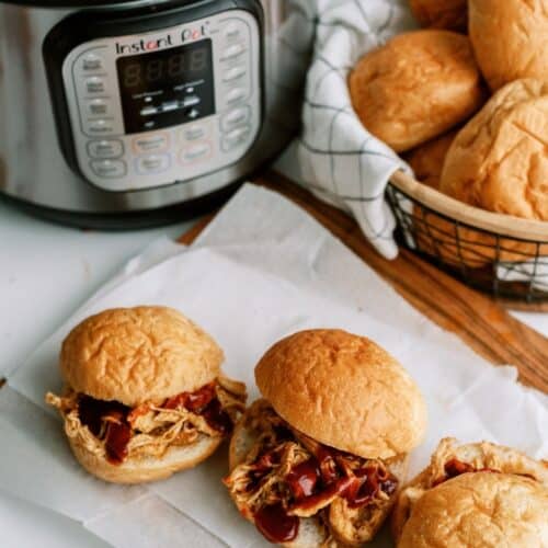 Three pulled chicken sandwiches with barbecue sauce on buns are on parchment paper next to an Instant Pot and a basket filled with more sandwich buns.