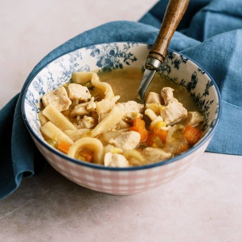 A bowl of chicken noodle soup with visible chunks of chicken, carrots, and noodles, with a spoon resting inside and a blue cloth underneath.