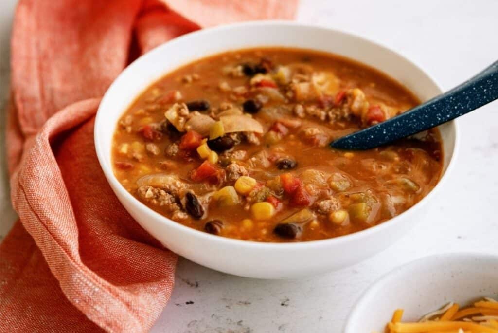 A bowl of hearty chili with beans, corn, tomatoes, and ground meat sits on a countertop next to an orange napkin and a bowl of shredded cheese.