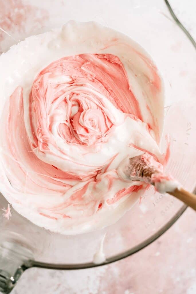 Marshamllows and cherry frosting mixed together in a glass bowl with a wooden spoon.