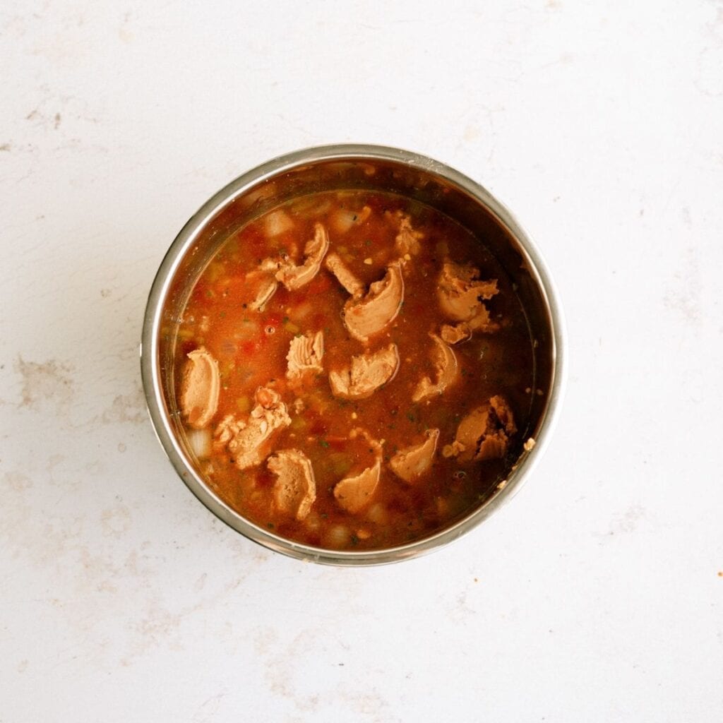 A metal bowl filled with broth containing chunks of meat and visible beans, photographed from above on a light surface.