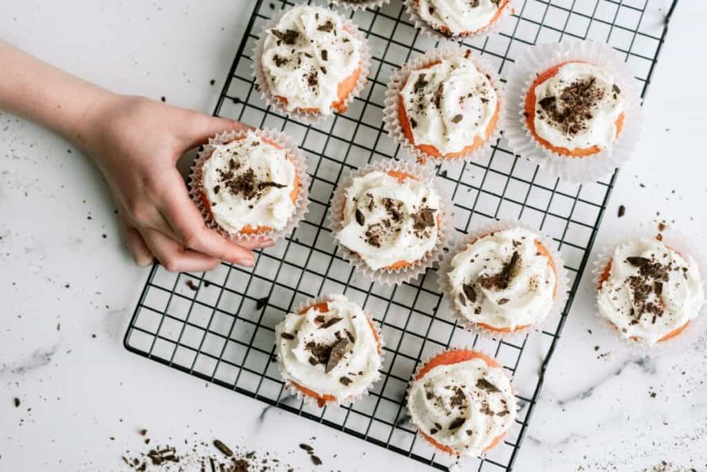A hand reaches for a cupcake topped with whipped cream and chocolate shavings on a wire cooling rack with scattered crumbs on a white surface.