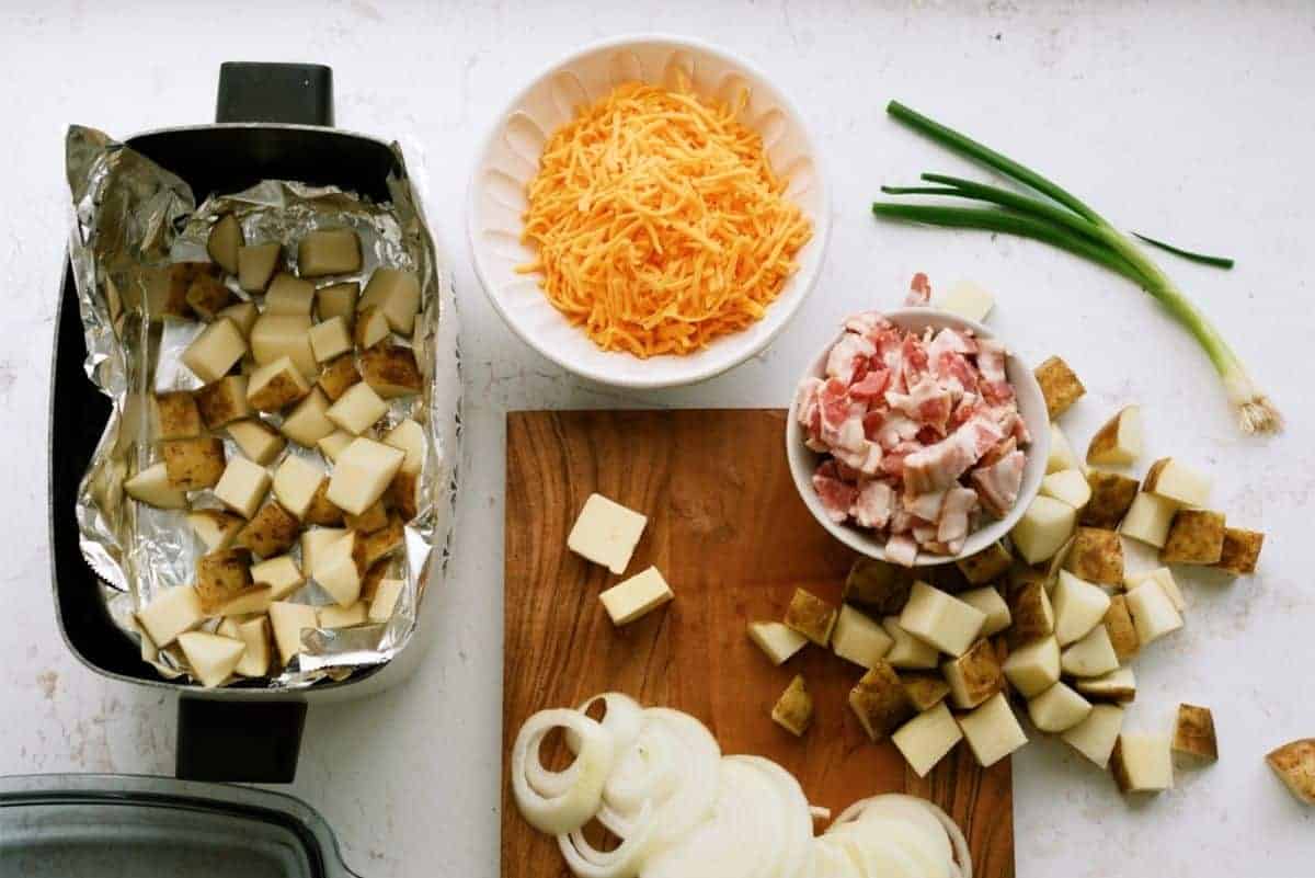 Chopped potatoes, bacon, and onions on a cutting board, shredded cheese in a bowl, and a baking dish lined with foil containing diced potatoes. Green onions are also on the table.