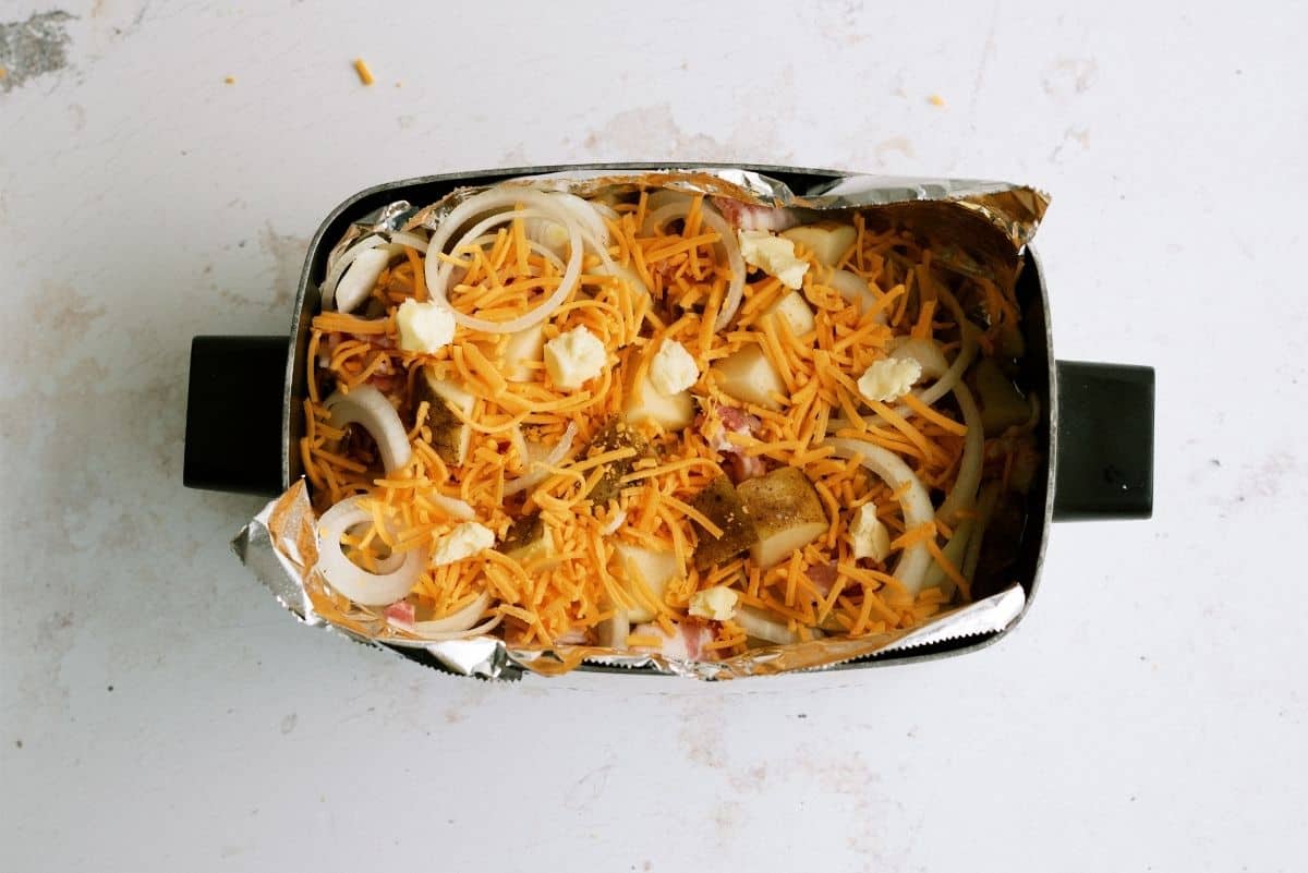 A rectangular baking dish lined with foil contains sliced onions, shredded cheddar cheese, and small chunks of butter on top of cut potatoes.