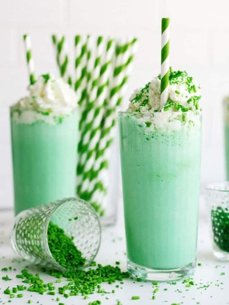 Two tall glasses of green milkshake with whipped cream and green sprinkles, each with a green-striped straw; a tipped glass spills green sprinkles in the foreground.