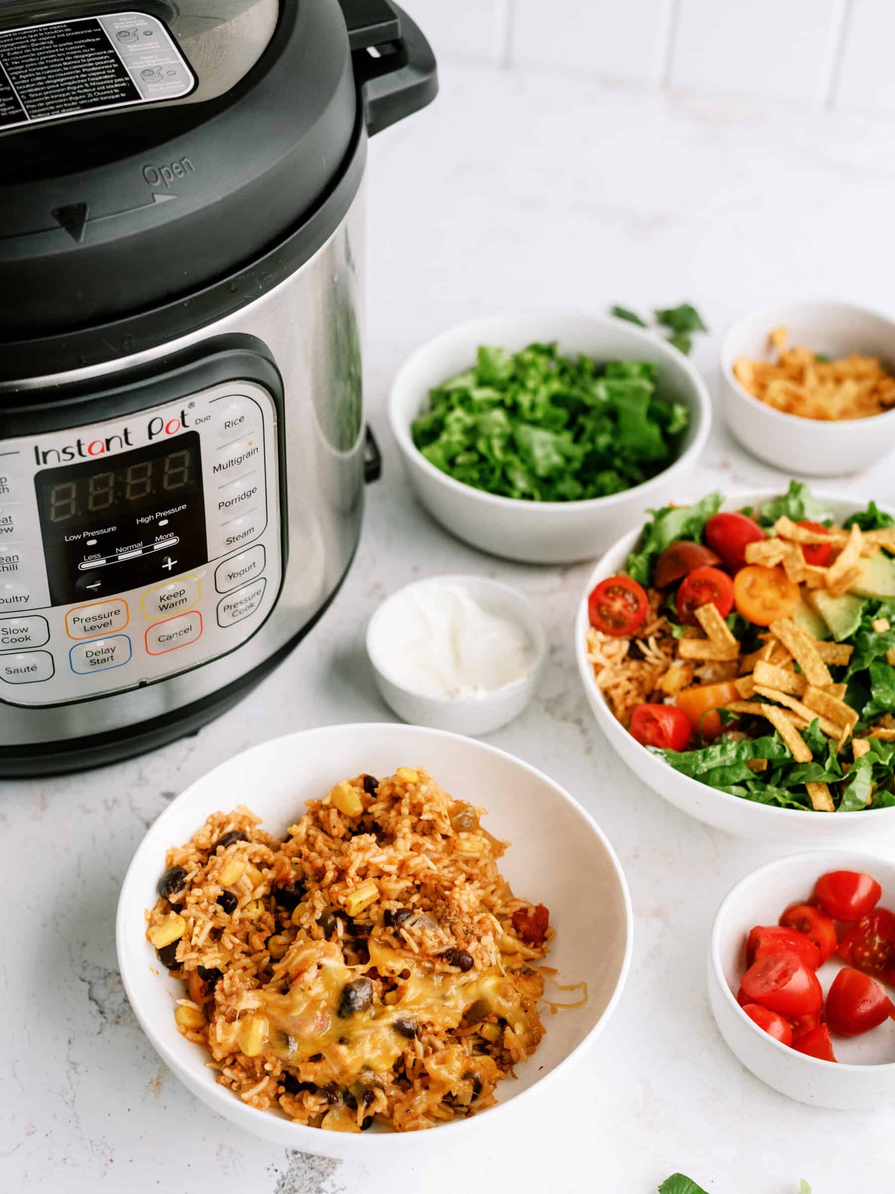 An Instant Pot surrounded by ingredients including one completely assembled burrito bowl, cheese, lettuce, sour cream, tomatoes, and a bowl of the ground turkey filling on a white countertop.