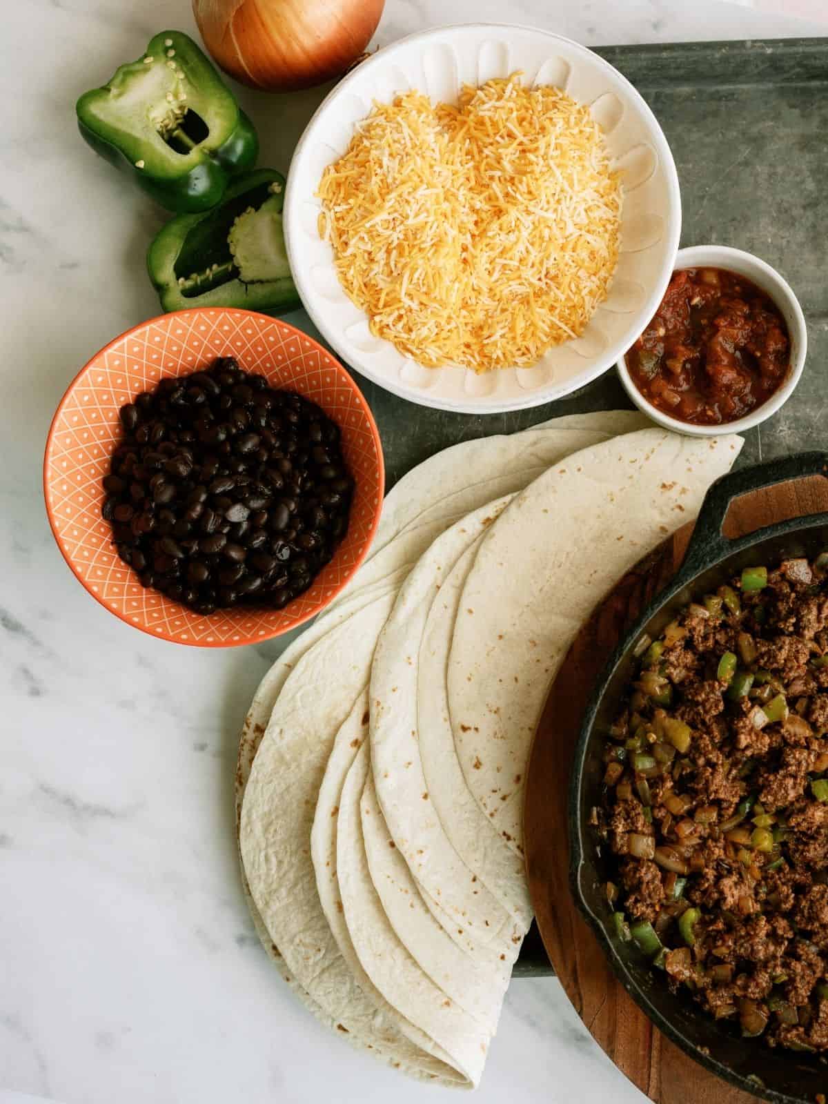 A spread of tortilla wraps, ground beef with peppers, black beans, shredded cheese, and salsa on a marble surface.