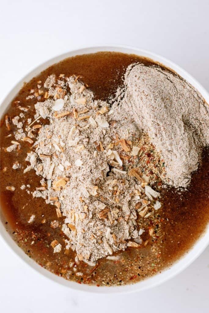 A bowl filled with layered dry and wet ingredients, including brown liquid, chopped nuts, and sandy textured powder.