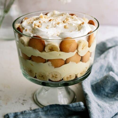 A glass trifle bowl filled with layered banana pudding, vanilla wafers, banana slices, and whipped cream, with a blue napkin beside it.