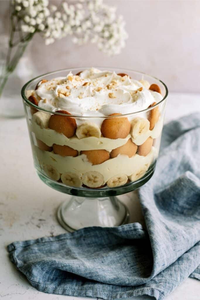 A glass trifle bowl filled with layered banana pudding, vanilla wafers, banana slices, and whipped cream, with a blue napkin beside it.