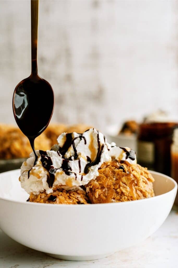 A spoon drizzling chocolate sauce on top of whip cream on fried ice cream in a bowl.