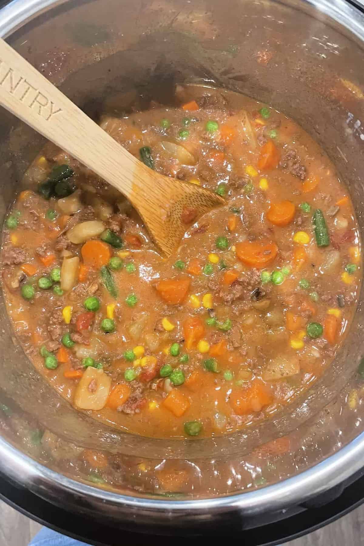 A wooden spoon stirring a pot of thick vegetable and meat stew with visible pieces of carrots, peas, corn, green beans, and potatoes.