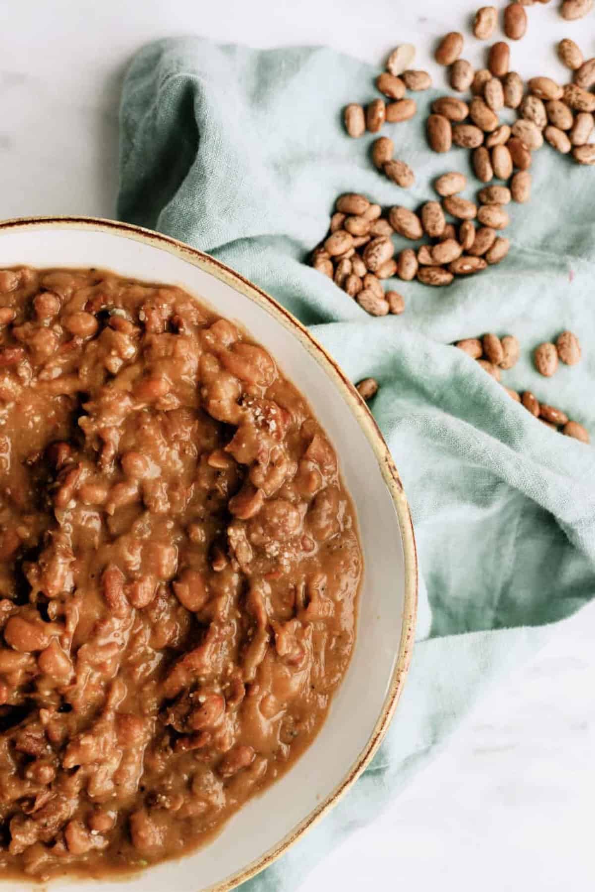 A bowl of refried beans sits on a light surface next to a green cloth with dried pinto beans scattered on top.