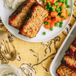 A plate with two slices of meatloaf, mashed potatoes, and mixed peas and carrots sits on a yellow floral tablecloth next to serving utensils and a casserole dish.