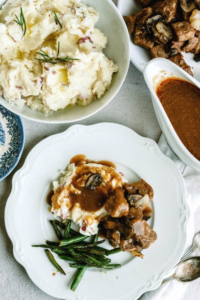 beef tips and gravy served over mashed potatoes on a white plate with green beans on the side, next to a bowl of mashed potatoes