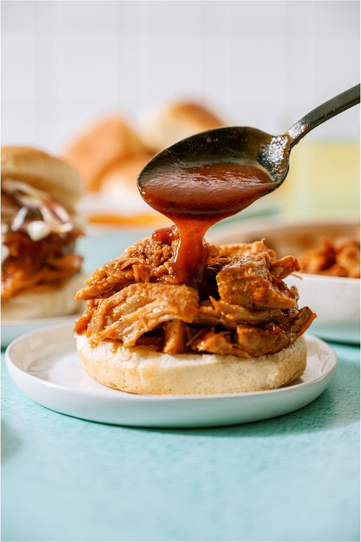 A spoon pouring BBQ sauce on top of shredded pulled pork on a bun.