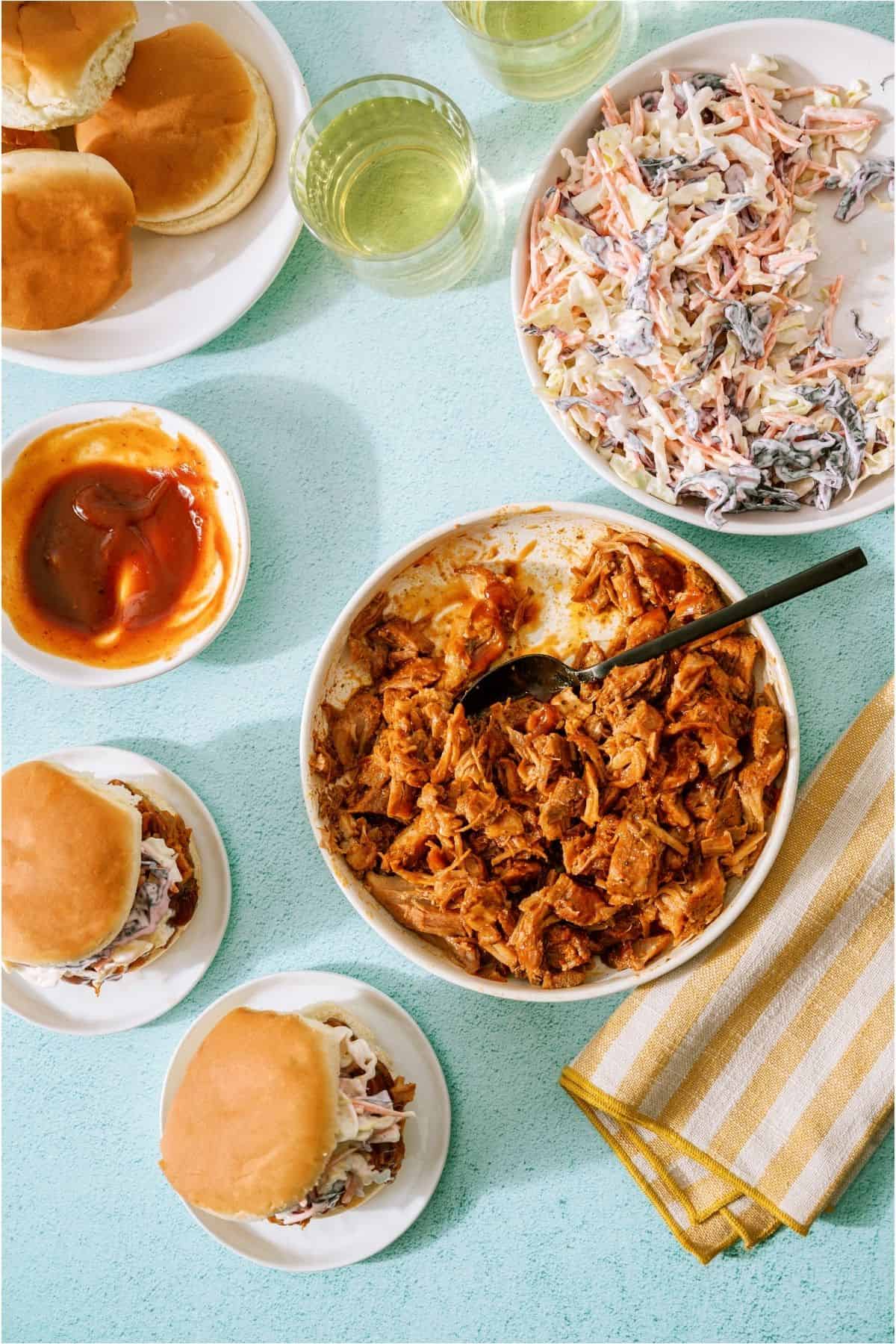 Top view of pulled pork sandwiches. A bowl of shredded pulled pork with a spoon in it and a bowl of coleslaw.