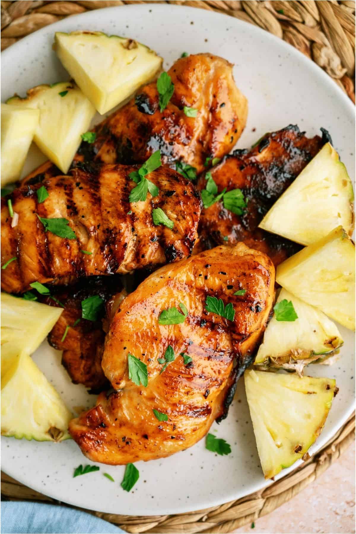 Top view of a plate of Grilled Huli Huli Chicken Breasts surrounded by slices of fresh pineapple.