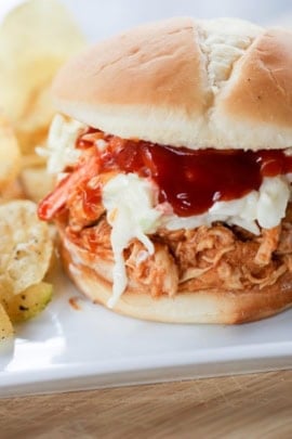 Close-up of a pulled chicken sandwich topped with coleslaw and barbecue sauce on a bun, with a side of potato chips.
