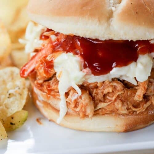 Close-up of a pulled chicken sandwich topped with coleslaw and barbecue sauce on a bun, with a side of potato chips.