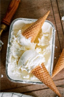 2 Ice Cream Cones with scoops on top of a container of Vanilla Ice Cream.