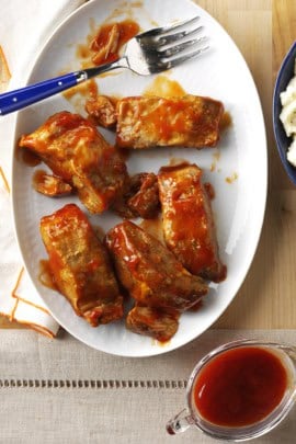 Plate of barbecue-glazed ribs with a fork, served next to a bowl of mashed potatoes and a pitcher of sauce on a wooden surface.