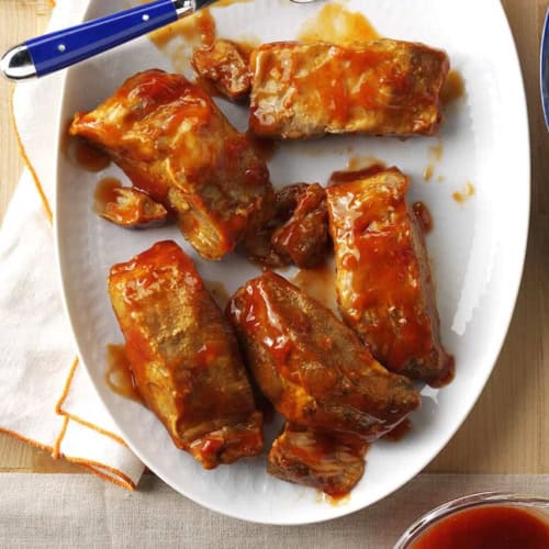 Plate of barbecue-glazed ribs with a fork, served next to a bowl of mashed potatoes and a pitcher of sauce on a wooden surface.