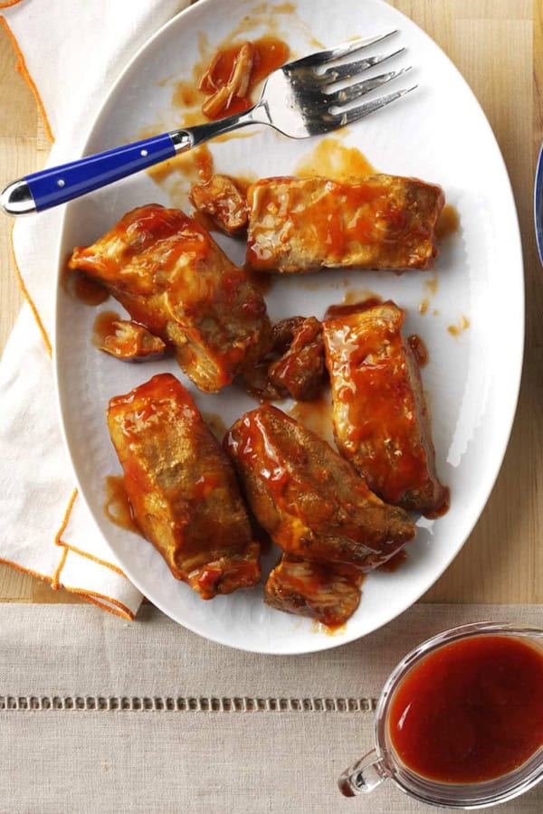 Plate of barbecue-glazed ribs with a fork, served next to a bowl of mashed potatoes and a pitcher of sauce on a wooden surface.