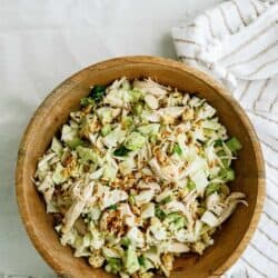 A wooden bowl filled with a chopped salad of cabbage, shredded chicken, sliced almonds, and noodles. A striped cloth lies beside the bowl.