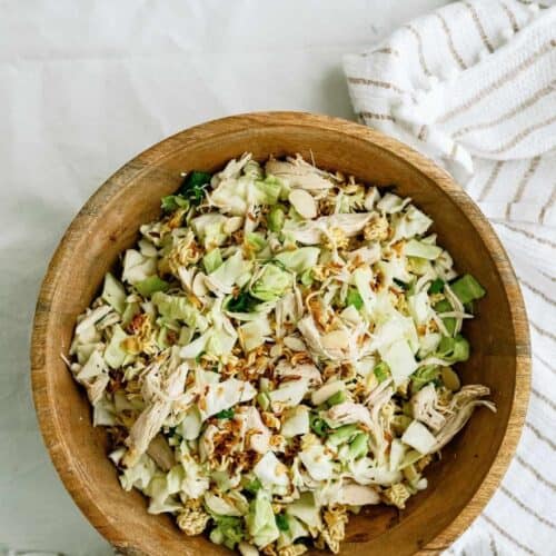 A wooden bowl filled with a chopped salad of cabbage, shredded chicken, sliced almonds, and noodles. A striped cloth lies beside the bowl.