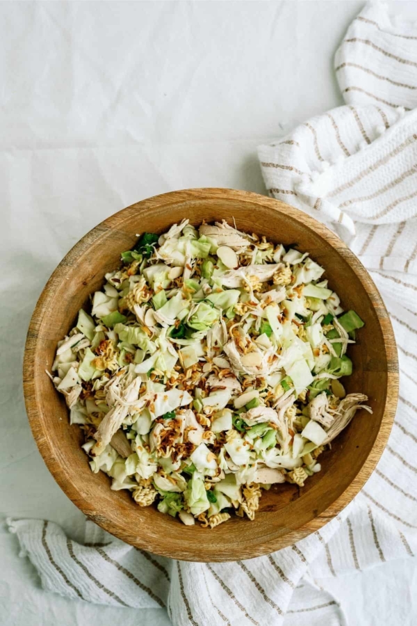 A wooden bowl filled with a chopped salad of cabbage, shredded chicken, sliced almonds, and noodles. A striped cloth lies beside the bowl.