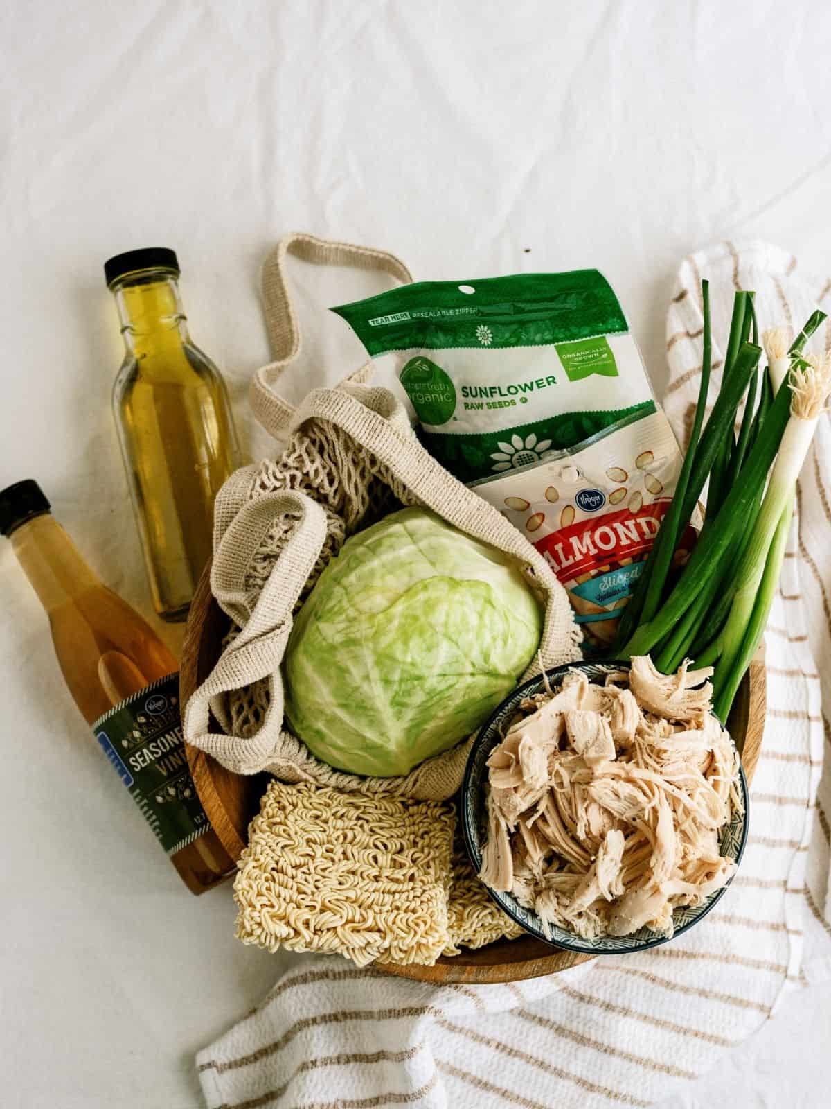 A bowl with cabbage, shredded chicken, green onions, ramen noodles, sesame dressing, sunflower seeds, and a bottle of oil on a striped cloth.