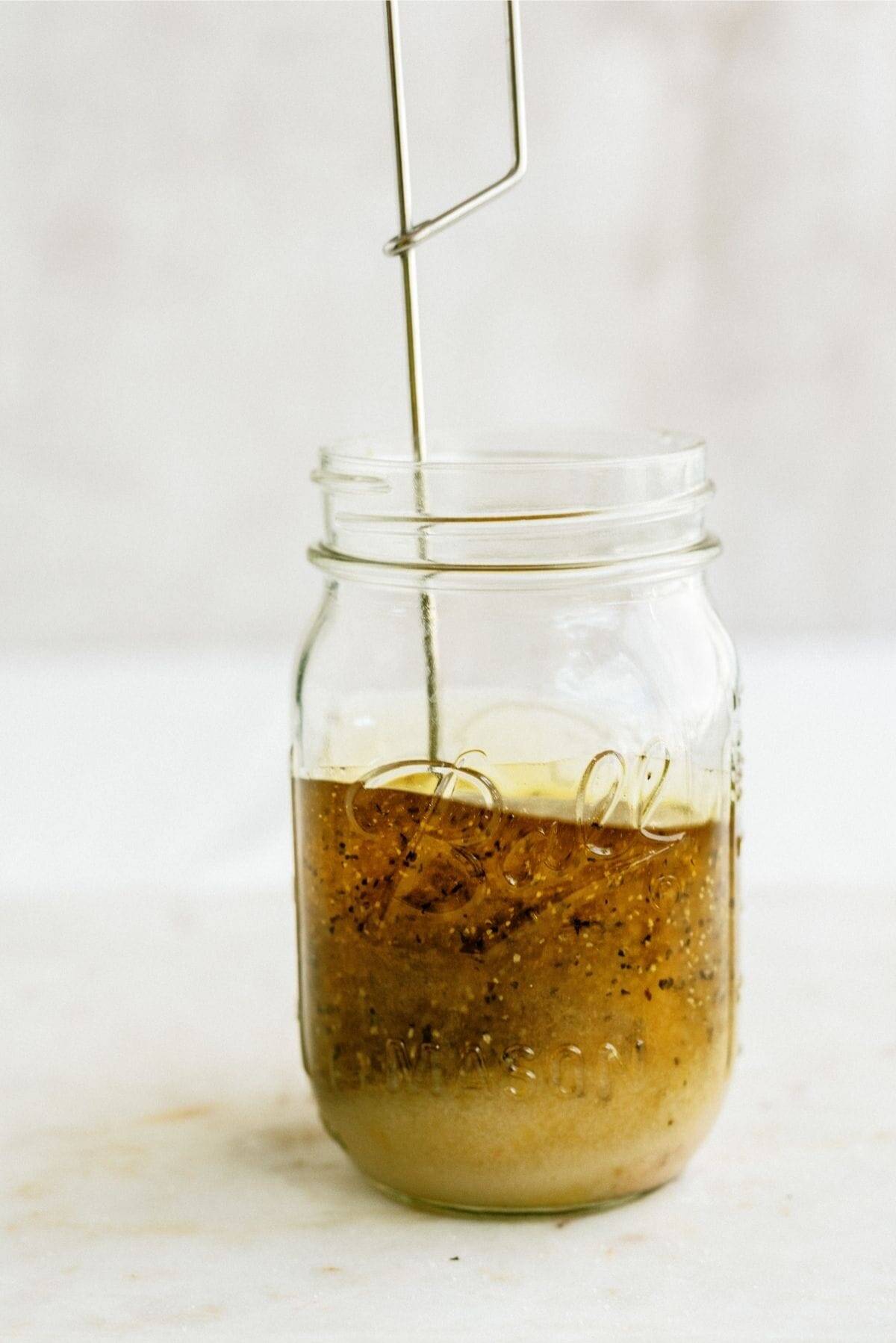 A glass jar filled with oil, vinegar, spices, and herbs, being stirred with a metal whisk.