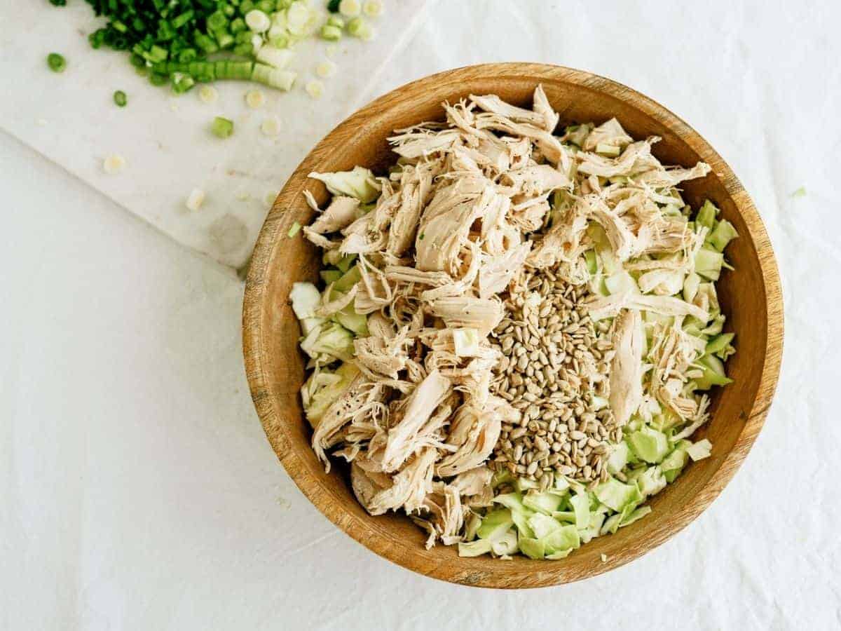 A wooden bowl filled with shredded chicken, chopped cabbage, and sunflower seeds on a white surface. Chopped green onions are on a cutting board in the background.