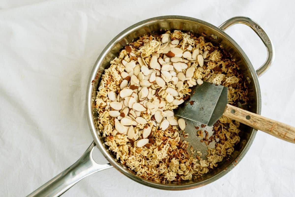 Pan with toasted ramen noodles, sliced almonds, and a wooden spatula on a white surface.