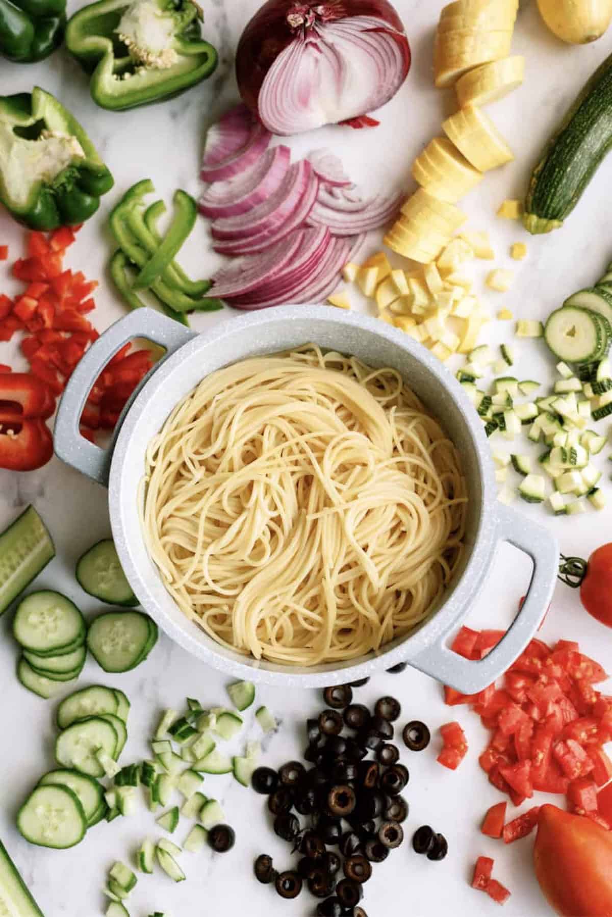A pot of cooked spaghetti is surrounded by sliced vegetables including cucumber, zucchini, olives, red onion, bell pepper, tomato, and yellow squash on a white surface.