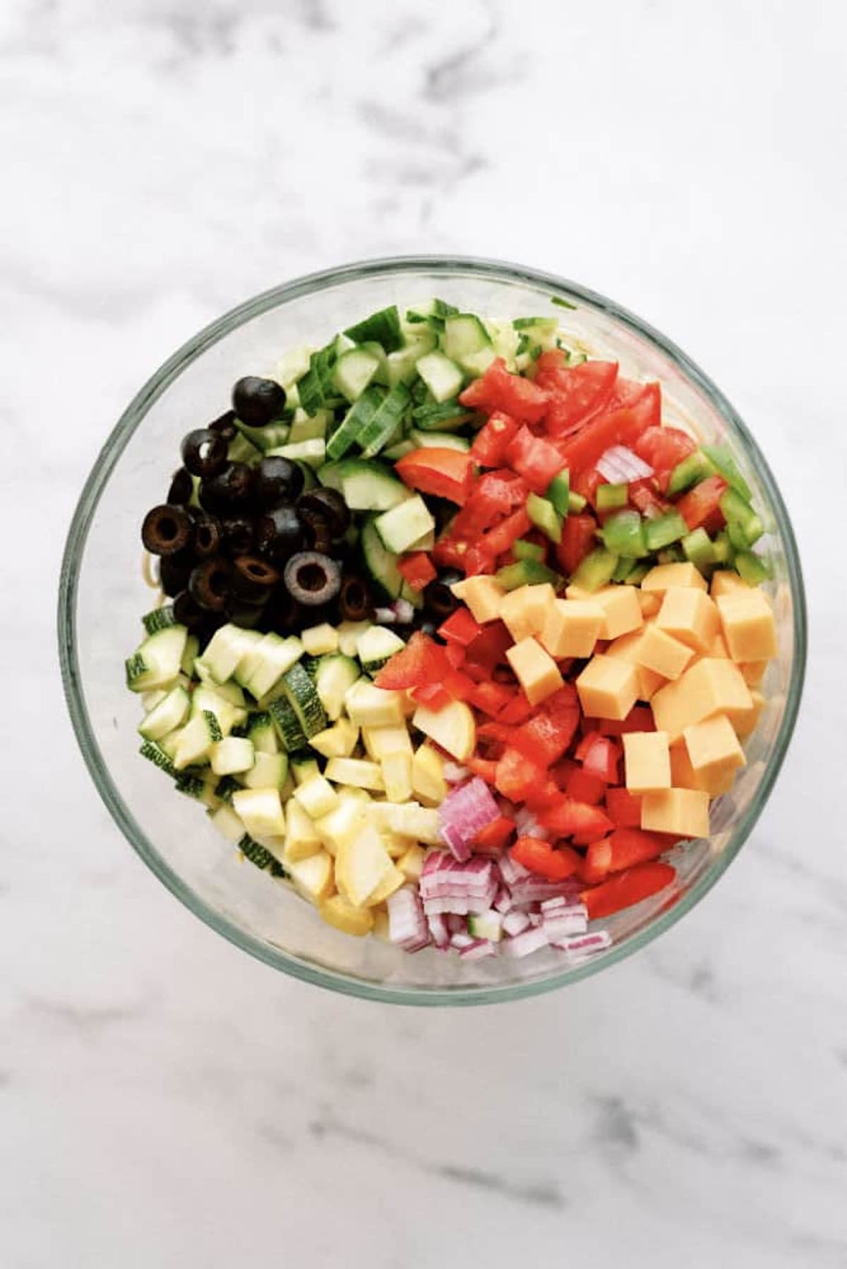 A glass bowl filled with chopped cucumber, tomatoes, green bell pepper, red onion, black olives, cheddar cheese cubes, and zucchini on a white surface.