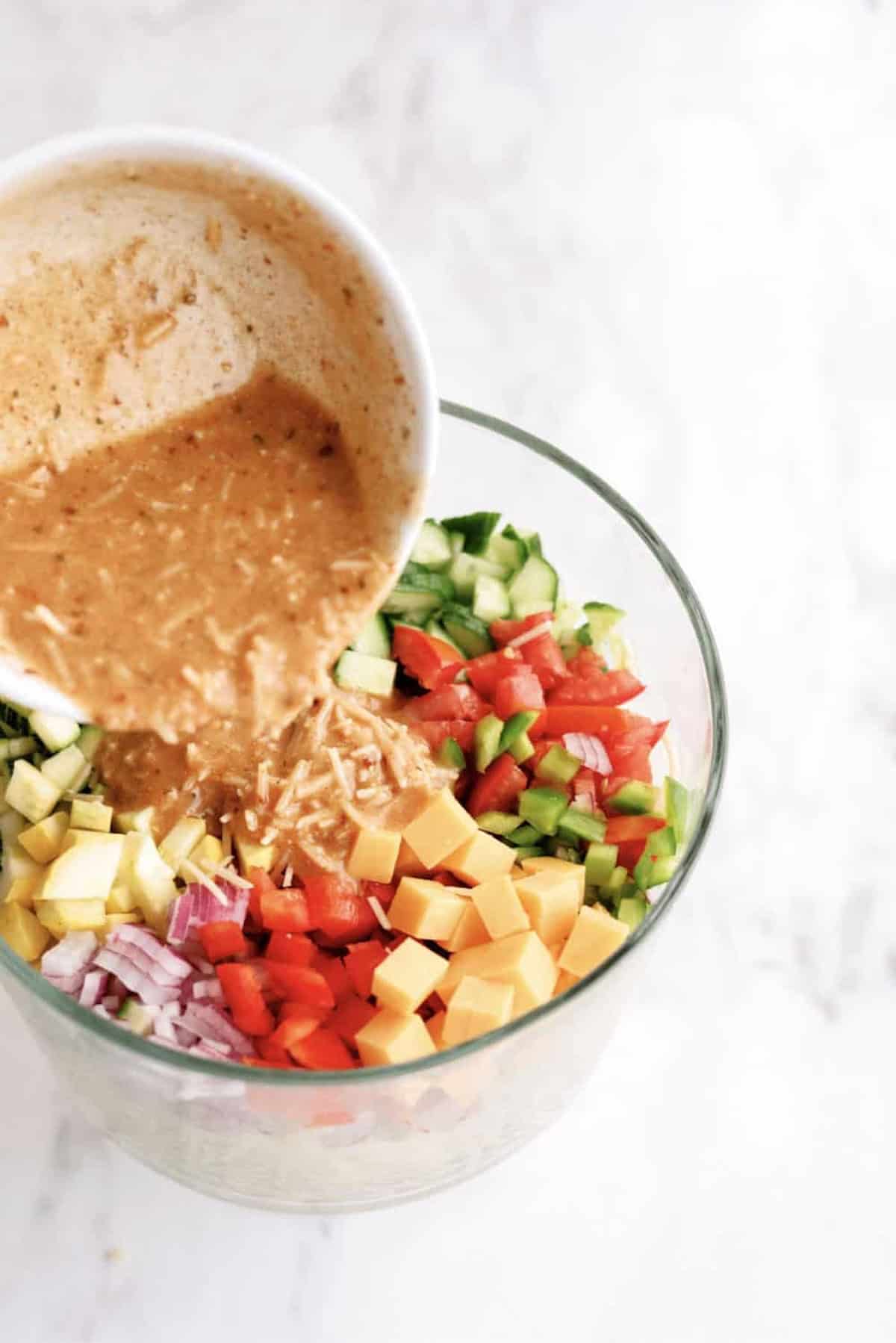 A glass bowl filled with chopped cucumber, tomatoes, green bell pepper, red onion, black olives, cheddar cheese cubes, and zucchini on a white surface.