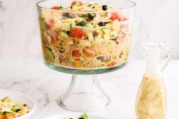 A glass trifle bowl filled with pasta salad containing vegetables sits next to a small pitcher of dressing and two smaller bowls of the same salad on a white surface.
