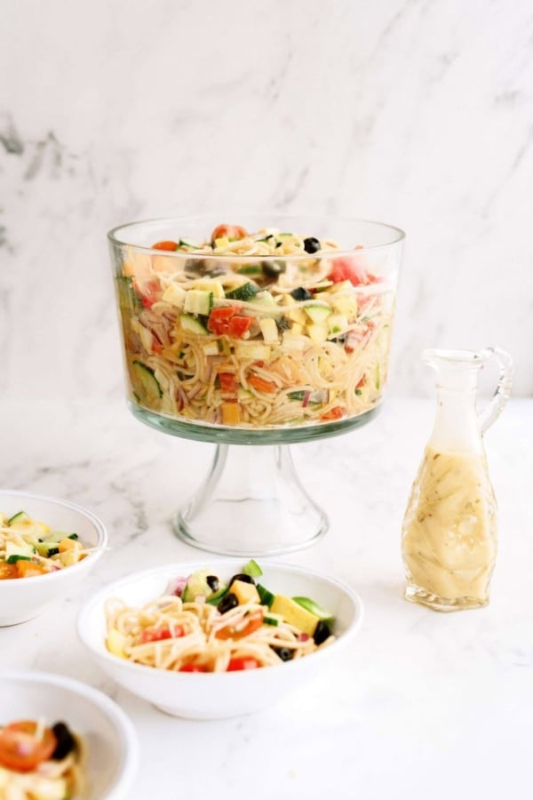 A glass trifle bowl filled with pasta salad containing vegetables sits next to a small pitcher of dressing and two smaller bowls of the same salad on a white surface.