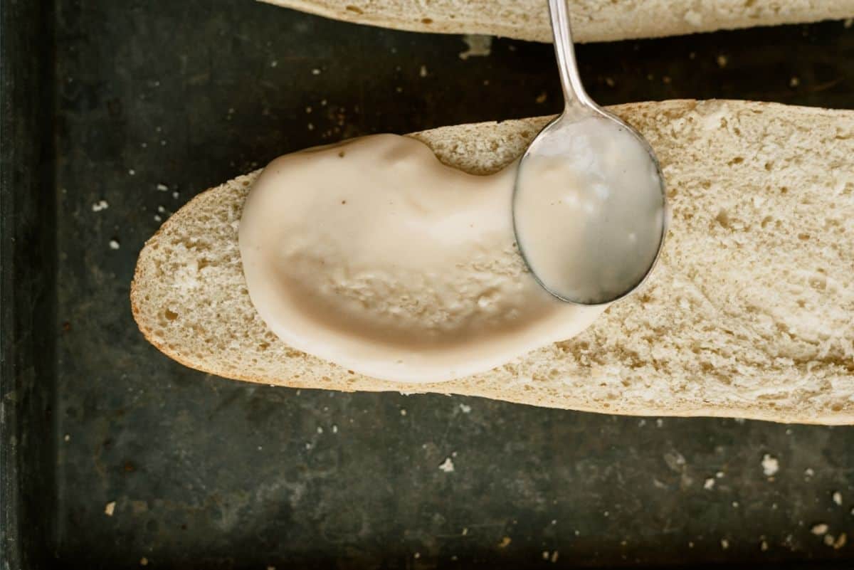 French Bread cut in half spreading Alfredo sauce onto each half with a spoon