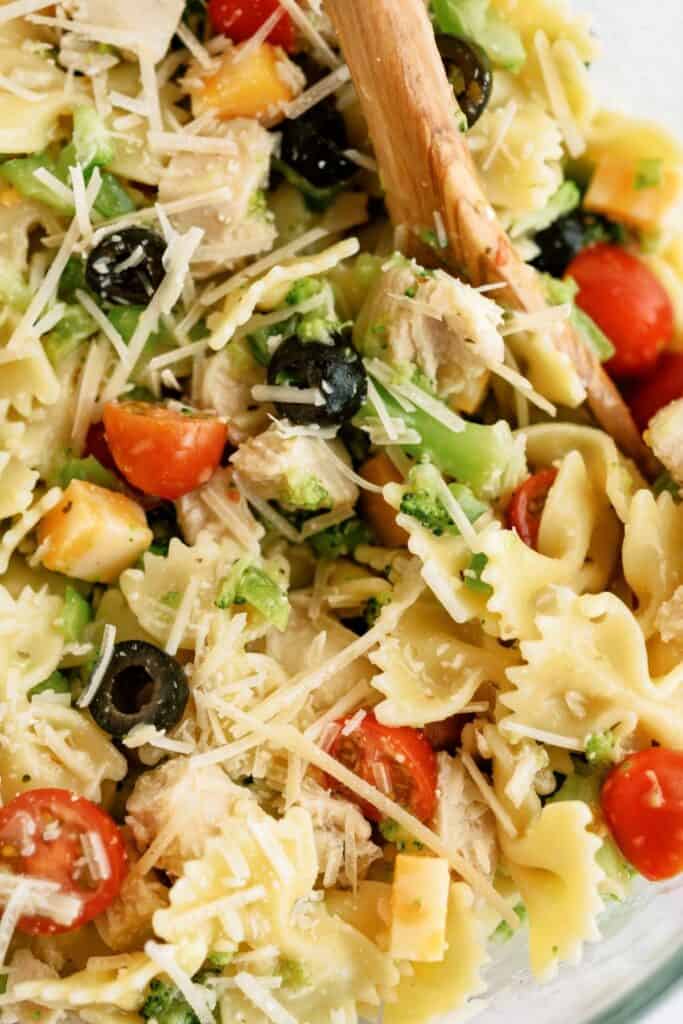 Close-up of a pasta salad featuring bow-tie pasta, cherry tomatoes, black olives, broccoli, diced cheese, and grated Parmesan, mixed with a wooden spoon.