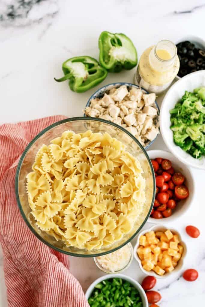Bowls of ingredients for pasta salad on a marble surface: uncooked bow tie pasta, green pepper, chicken, dressing, black olives, broccoli, tomatoes, cheese cubes, green onions, and a red cloth.