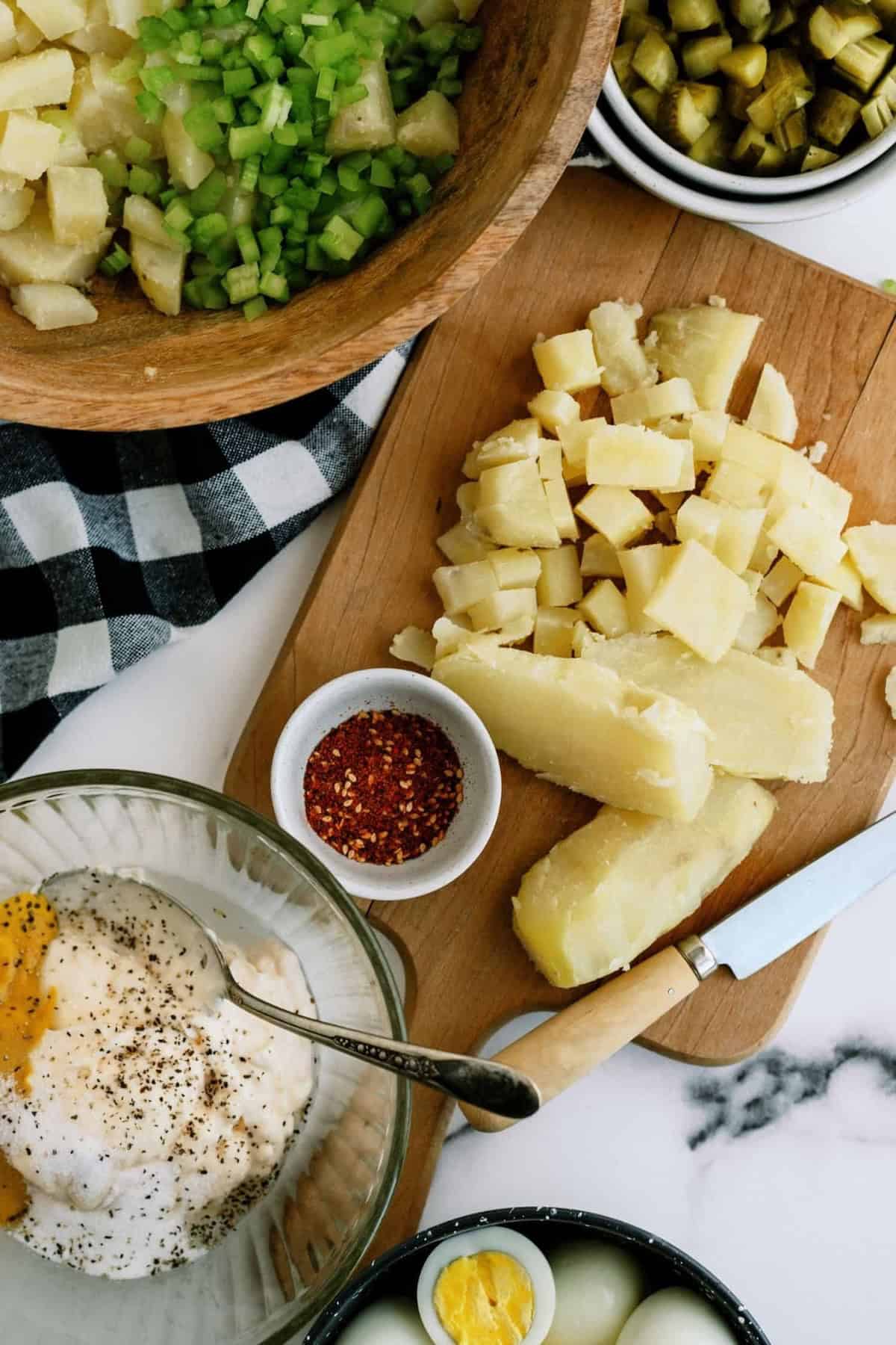 Chopped potatoes, celery, pickles, and a spice mix are arranged on a board, with a knife beside them. A bowl with mayonnaise and hard-boiled egg slices is also visible.