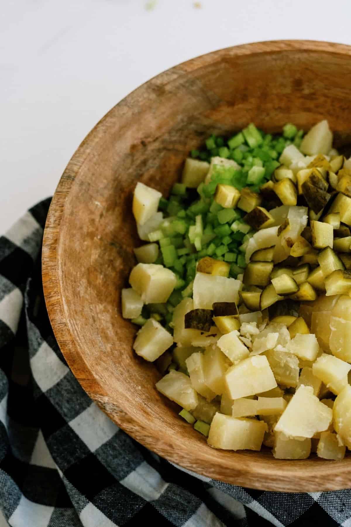 Wooden bowl with chopped potatoes, celery, and pickles on a black and white checkered cloth.