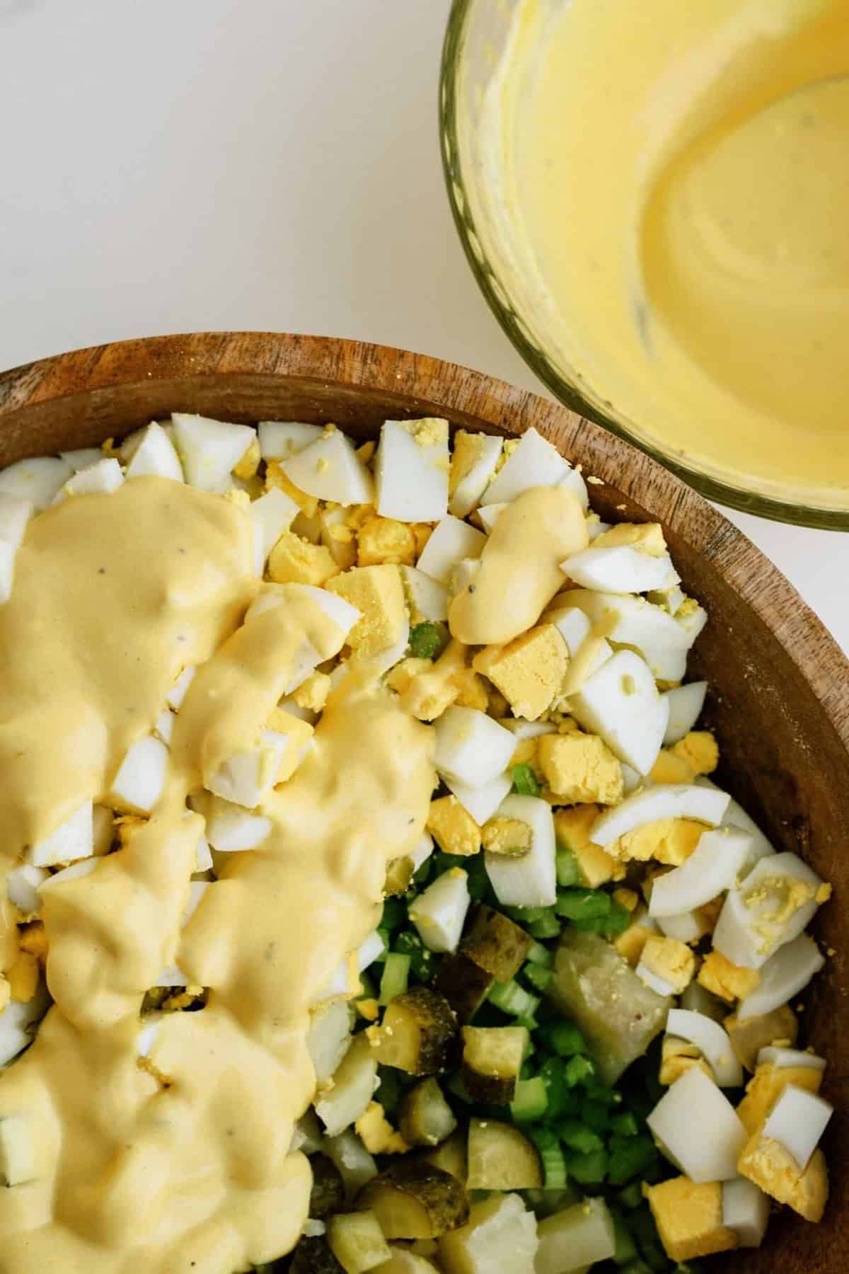 Chopped boiled eggs, diced vegetables, and dressing on a salad in a wooden bowl; extra dressing in a glass bowl beside it.