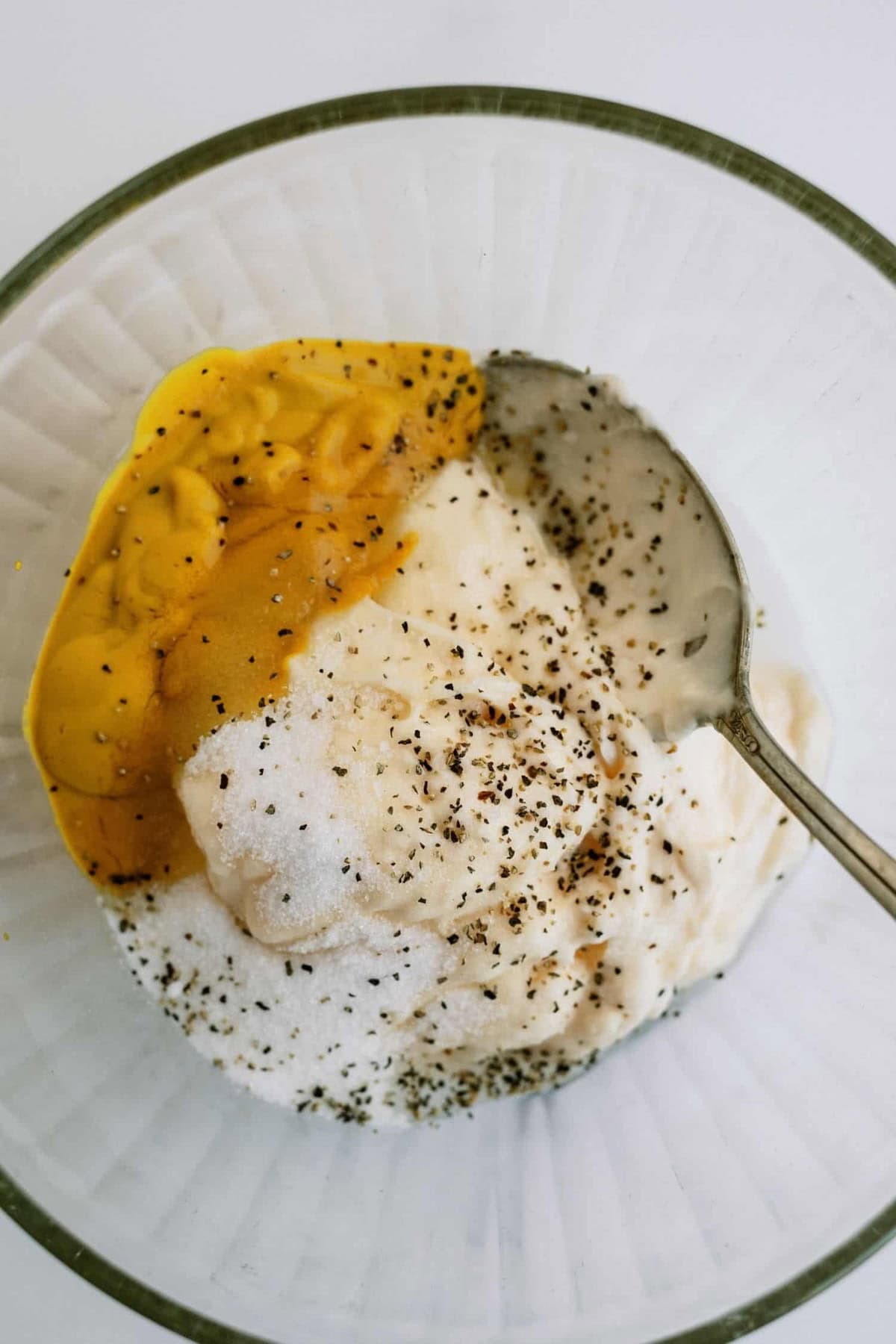 A bowl containing mayonnaise, mustard, sugar, salt, pepper, and a spoon.